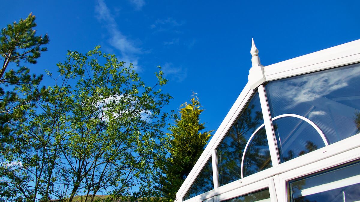 cropped image showing apex of white upvc conservatory against bright blue skies and a big green-leafed tree